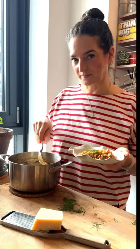 A woman wearing a white and red striped top looks at the camera whilst holding a plate with one hand and dipping a spoon in a pot with the other.