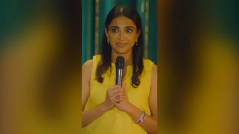Sindhu Vee wearing a yellow top and holding a mic.