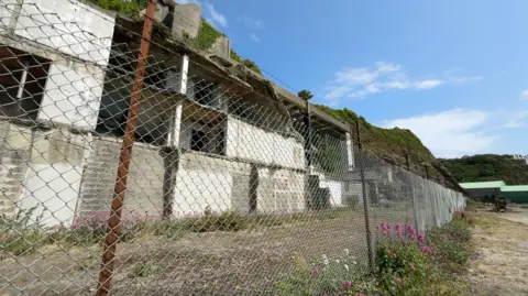 The derelict site, a high metal fence runs along it, you can see the remaining parts of a building structure in front of a cliff. 