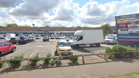 A shopping centre car park. There are several cars parked and a white van is driving through the car park. On the right is a sign with the words "Staple Tye Shopping Centre" above the logos of 18 businesses.