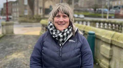 BBC A blonde haired woman with a black and white scarf and blue jacket smiles at the camera