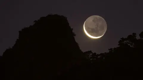 A thin crescent Moon with the darker whole visible, low on the horizon