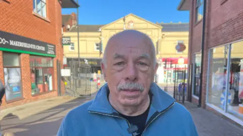 Stan Crook wears a blue jacket. He is looking at the camera. He has a moustache.