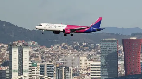 A white, pink and blue airplane with "Wizz" written on the side flies low over a city, with wooded hills in the background.