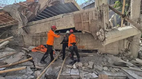 EPA Rescuers clad in bright orange long sleeved vests and helmets inspect the rubble of a collapsed building at an Islamic boarding school in Sidoarjo, East Java, Indonesia. They are standing in front of a huge heap of twisted concrete and rebar.