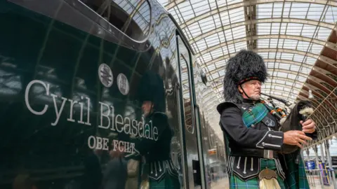 A piper in green and back tartan Highland Dress performs on the platform alongside the Great Western Railway train named after Cyril Bleasdale OBE.