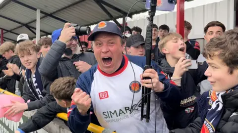 Connor Bennett/BBC James Beardwell wearing a white and blue strip and blue baseball cap holding a long phone stand in his left hand and raising his clenched right fist while roaring enthusiasm. He is leaning over the stand surrounded by mostly young fans. 