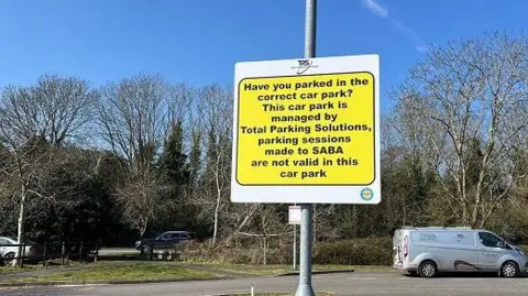 Total Parking Solutions Sign in black writing on yellow background telling drivers Saba payments are "not valid in this car park".  There is a car park behind in which a white van is parked. There are trees beyond the car park.