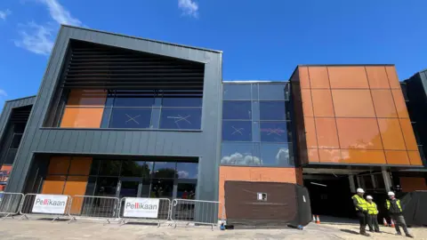 A large modern building with external walls that are a mixture of copper-coloured and dark material is seen from the outside on a sunny day. At the foot of one of the walls are three people wearing yellow high-viz jackets and white hard hats