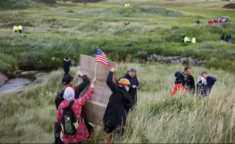 EPA Three of protesters holding an American flag and a placard in the grounds of Turnberry with their backs to the camera. They are standing in a tuft of long grass with a golf course stretching in front of them. The resort's buildings can be seen in the background.