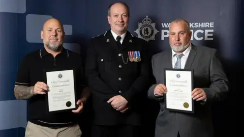 Warwickshire Police Three people stand in front of a Warwickshire Police backdrop. A senior police officer in full dress uniform, wearing medals, stands in the centre. On each side, a person holds a framed ‘Chief Constable Commendation’ certificate, smiling as they pose for the photo