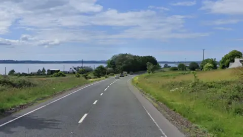 Google A single carriageway road with grass on either side and a river can be seen in the distance