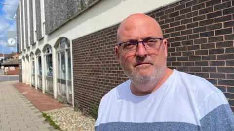 Andrew Turner/BBC Ian Measures is wearing a blue top, with lighter and darker shades in horizontal bands. He has a shaved head, a close cropped beard and moustache, and is wearing glasses. He is standing by Gorleston Library. The building is a 1970s design, featuring brown bricks, concrete pillars and arches, several infilled with aluminium-framed windows, and flint-faced concrete panels on the upper storey.