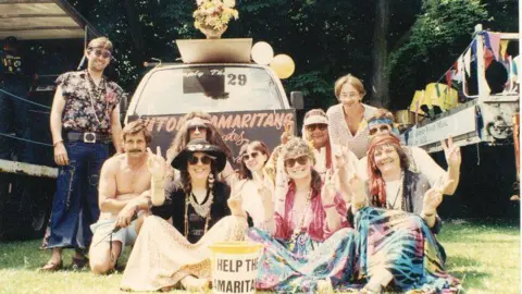 Matthew Catlin A group of people dressed in clothing reminiscent of the "Summer of Love" pose in front of a car with the words "Luton Samaritans" written on it