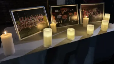 PA Media Three framed pictures are sitting on a table with candles. The picture in the centre is a composite of five portraits of young people.