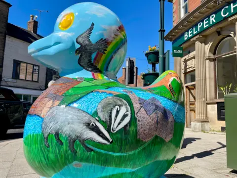 A giant duck sculpture sits outside a former bank building. The duck has a detailed drawing of badgers, a fox and birds over a countryside view of a mill beside a river. It's eyes are yellow.