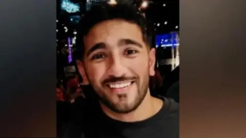 Rehaan Akhtar smiling. He is wearing a black T-shirt. Behind him, what appears to be a nightclub can be seen.