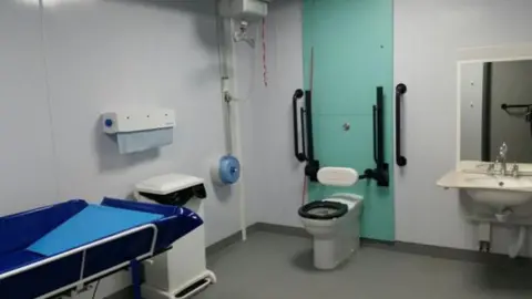 An accessible changing place facility, with a hoist, a bed, toilet and sink. It is a white room with black hand rails to improve accessibility.