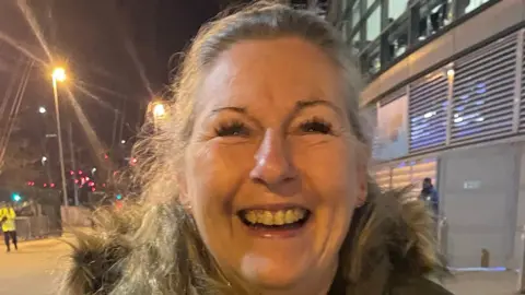 Plymouth Argyle fan Jacqueline smiles as she leaves the Etihad Stadium, which is to her right. It is night time and a police officer wearing a yellow high-vis coat is walking behind her.