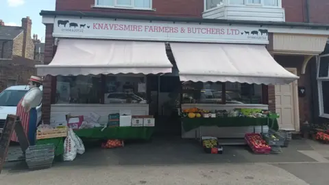 The exterior of a traditional butchers shop, with a white fabric awning and an outdoor display of fruit and vegetables. 