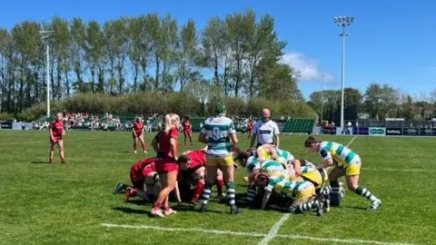 Two women's rugby teams - one in red and one in green and white hoops - set up for a scrum, with a linesman and referee watching.