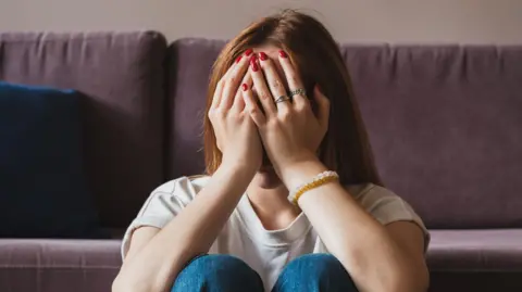 A woman holds her hands over her face. She is sitting on a floor with a purple couch in the background.