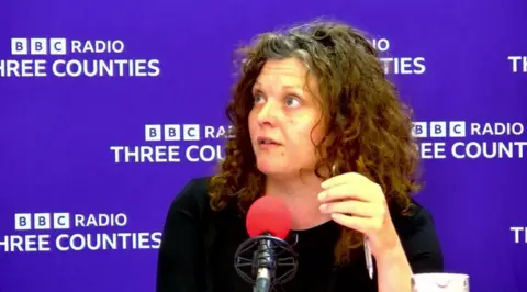 Zara Layne with long, brown curly hair sits in a radio studio behind a microphone with a pink windscreen. The backdrop displays repeated BBC Radio Three Counties branding.