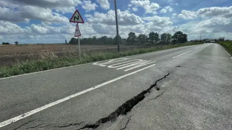 A large crack in a tarmac road in a rural location, with fields surrounding it and blue skies overhead.
