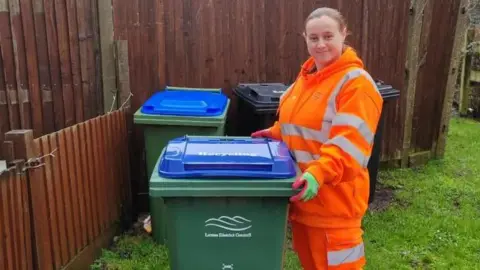 A woman with long hair wearing orange high vis trousers and jacket. She is standing next to a bin which says "Lewes District Council" on it.