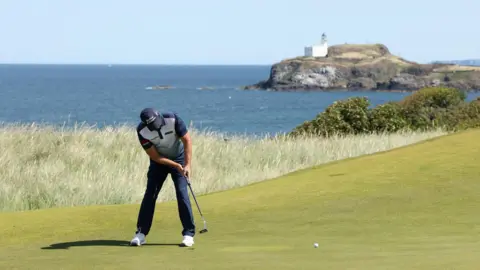 Getty Images Padraig Harrington rolls a putt on the 13th green during the pro-am. He is wearing a navy cap, white and navy polo shirt, navy trousers and white golf shoes. A white lighthouse is visible on a rock in the background.