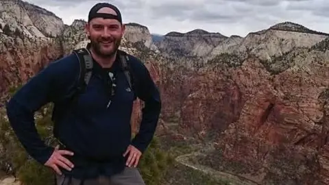 A person wearing a dark long-sleeve top, a backpack, and a backwards cap stands on a mountain overlook with hands on hips. Behind them, steep red and beige rock formations stretch across a canyon landscape under a cloudy sky.