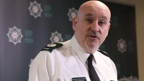 PA Media A man with a white stubble beard wearing a white shirt with dark green shoulder badges and tie