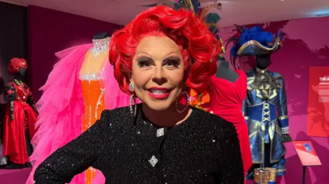 La Voix is standing in front of some costumes from the exhibition. She has short, bright red hair, and is wearing a black sparkly top, with silver diamond buttons, and silver hoop earrings. Behind her are an array of her costumes, all brightly coloured. There is a bright pink and orange one to her left, and a Captain Cook inspired drag costume to her right.