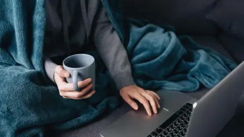 A man sits with a blanket around him and cups a hot drink as he sits at a laptop.
