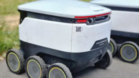 A small delivery robot. It is white and grey and less than a meter high and has six wheels. It is parked next to another robot.