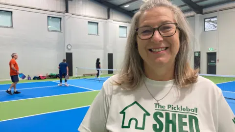 BBC/Julie Mariotti Gill Page is facing the camera and smiling with a white T-shirt on that says The Pickleball Shed. She has blonde hair and dark rimmed glasses. Behind her, you can see the pickleball court which has a blue and green flooring with three people holding rackets amid a game of pickleball. 