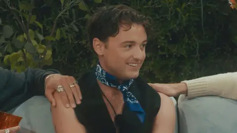 Adam is wearing a black vest and dark blue neck tie. Two people, who are out of shot, have their hands resting on his shoulder.