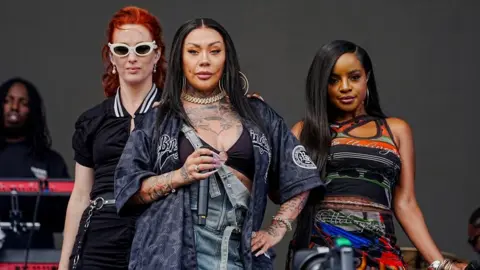 The Sugarbabes perform on a stage. A lead performer stands at the centre, holding a microphone mid‑song. She is wearing a loose, dark sports-style top and dungarees. Two other performers stand slightly behind and to each side. Musical equipment, including a keyboard, is visible in the background.