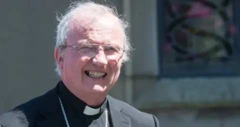 Donal McKeown is smiling at the camera. He is wearing a black jacket white and clerical collar with a large silver chain around his neck. He has white hair and gold glasses