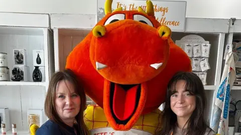 Devon County Show Two women with dark long hair are stood either side of a red dragon mascot that is wearing a Pete's Dragons t-shirt