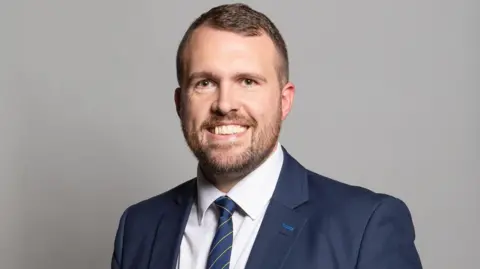 Jonathan Gullis - a man with brown hair, a beard, a blue suit jacket, blue striped tie and white shirt - smiles while in front of a grey wall.