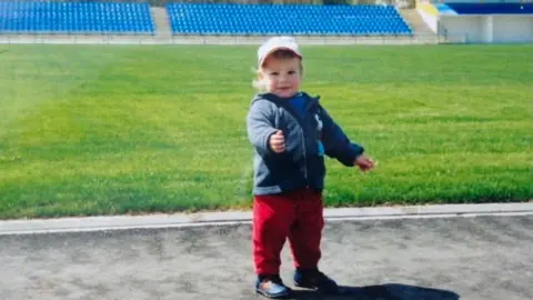 Ivan Varfolomeev A young child wearing red trousers and a blue jacket, standing inside an empty sports stadium.