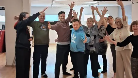 Tang Hall SMART CIC A mixed gender group of older people raise their arms in the air. In the centre, a younger man with short brown hair and a beard stands next to a young man with Down's syndrome. The group stand in a white studio space, with wooden flooring.