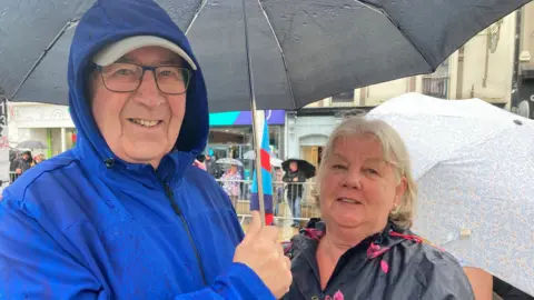 Shirley Henry / BBC Roger and Sheila Harvey stand beneath an umbrella. He wears glasses and a blue rain jacket. She has shoulder-length blond hair and a black rain jacket with red flowers on 