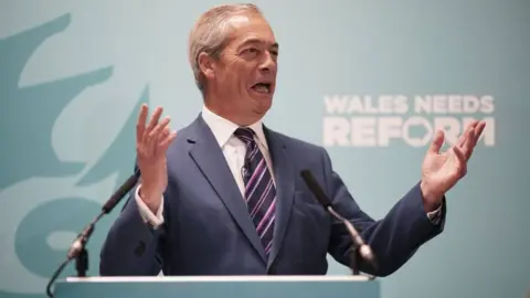 PA Media Nigel Farage speaking at a podium in front of a board that reads 'Wales needs reform'