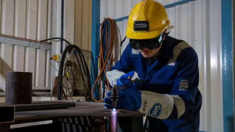 Handout A man in working gear with protective glasses and a helmet is using what appears to be a blow torch or a welder 