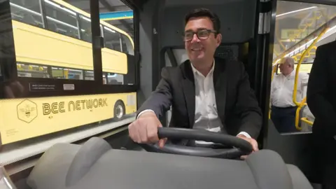 PA Media Andy Burnham drives a yellow "Bee Network" branded bus.