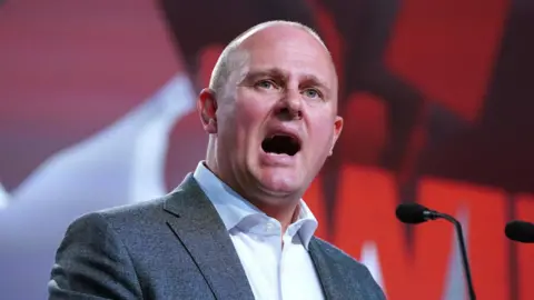 PA Media Paul Nowak, wearing a grey suit and white shirt, speaks from a podium with a red wall behind him. 
