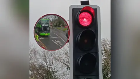 A trixi mirror is attached to a traffic light. A green bus is seen in the reflection of the round mirror alongside a red traffic light.