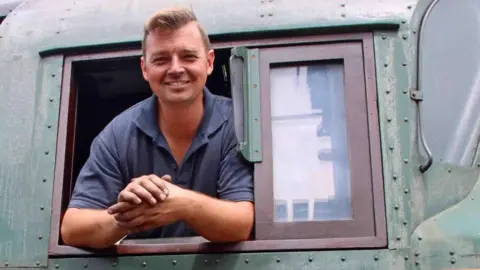 Andrew PM Wright A smiling Billy Johnson leaning out of the window of a locomotive. He has short blond hair and is wearing a blue polo shirt.